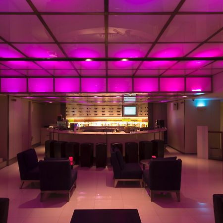 Pathumwan Princess Hotel - Sha Extra Plus Certified Banguecoque Exterior foto The photo shows a modern bar interior with a sleek design. The lighting features a pink hue that illuminates the ceiling, creating a vibrant atmosphere. In the center, there is a rounded bar counter with a well-stocked shelf behind it. The seating ar