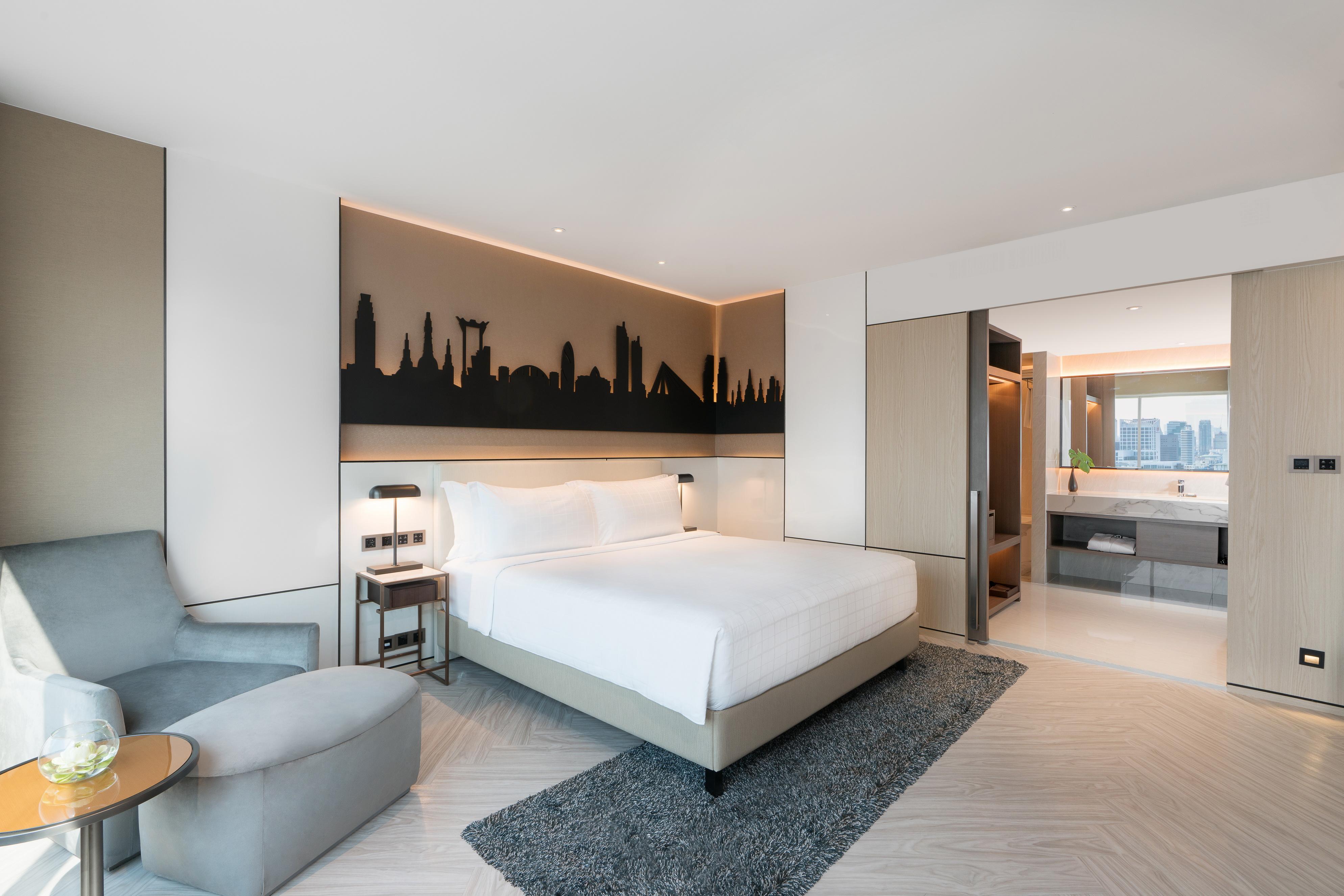Pathumwan Princess Hotel - Sha Extra Plus Certified Banguecoque Exterior foto The photo shows a modern hotel room featuring a minimalistic design. There is a large, white bed with a headboard that has a silhouette of a city skyline illuminated by soft lighting. To the left, there is a comfortable gray armchair next to a small 