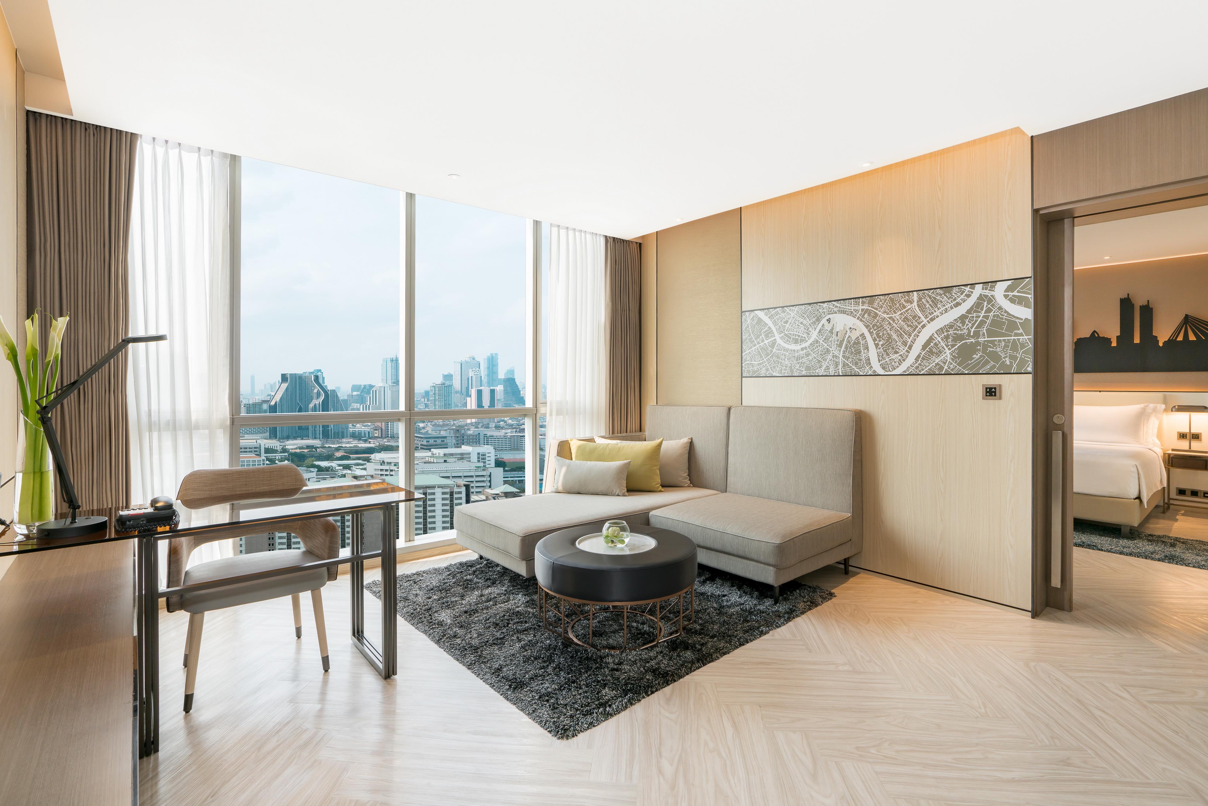 Pathumwan Princess Hotel - Sha Extra Plus Certified Banguecoque Exterior foto The photo shows a modern, stylish living area in a hotel or apartment. It features a large window that offers a view of a city skyline. The room is designed with neutral-colored walls and light wooden flooring. There is a comfortable gray sofa with t