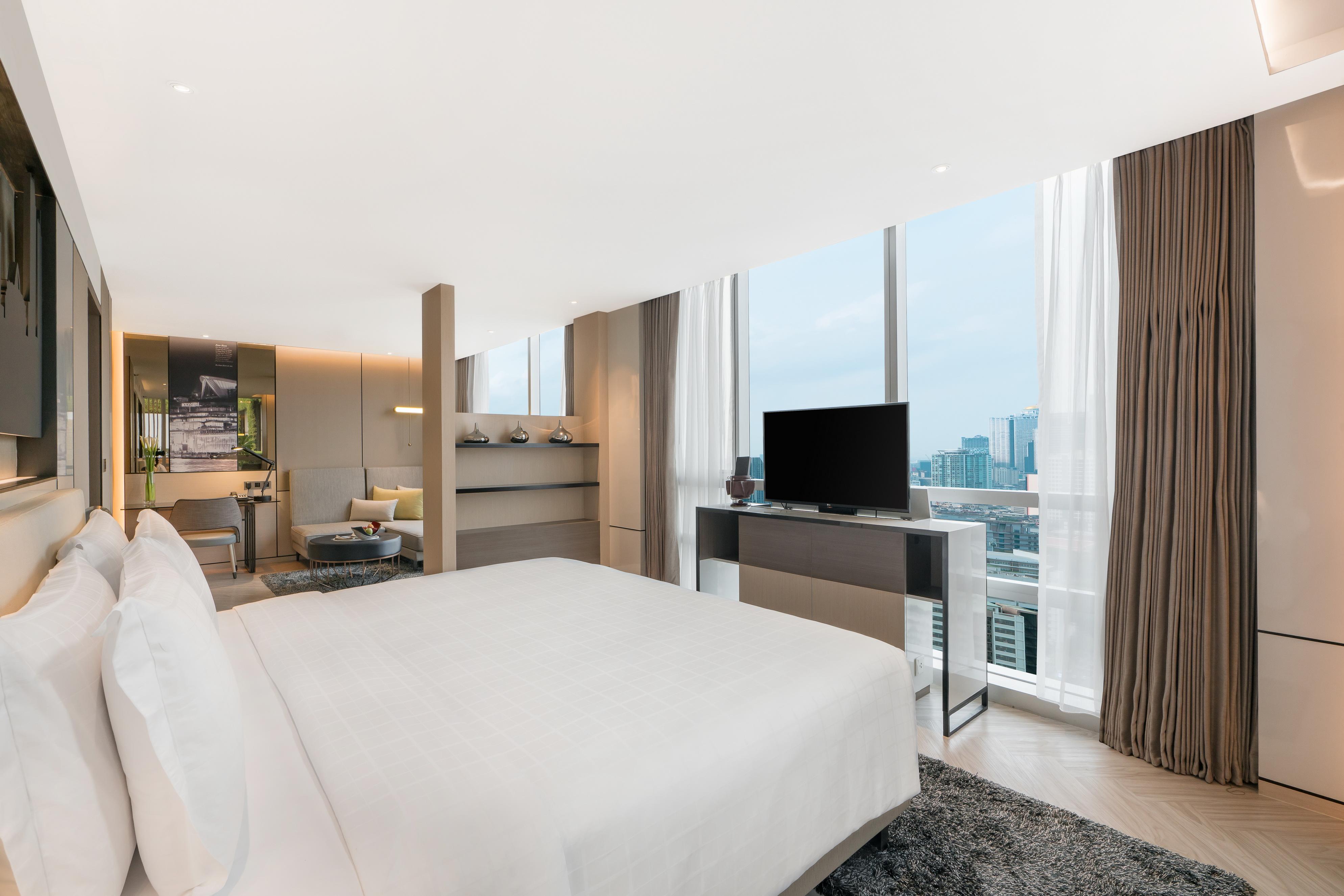 Pathumwan Princess Hotel - Sha Extra Plus Certified Banguecoque Exterior foto The photo shows a modern hotel room, featuring large windows that provide a view of the city. The room has a spacious layout with a king-sized bed covered in white linens, a bedside table, and a contemporary television mounted on a console. There are