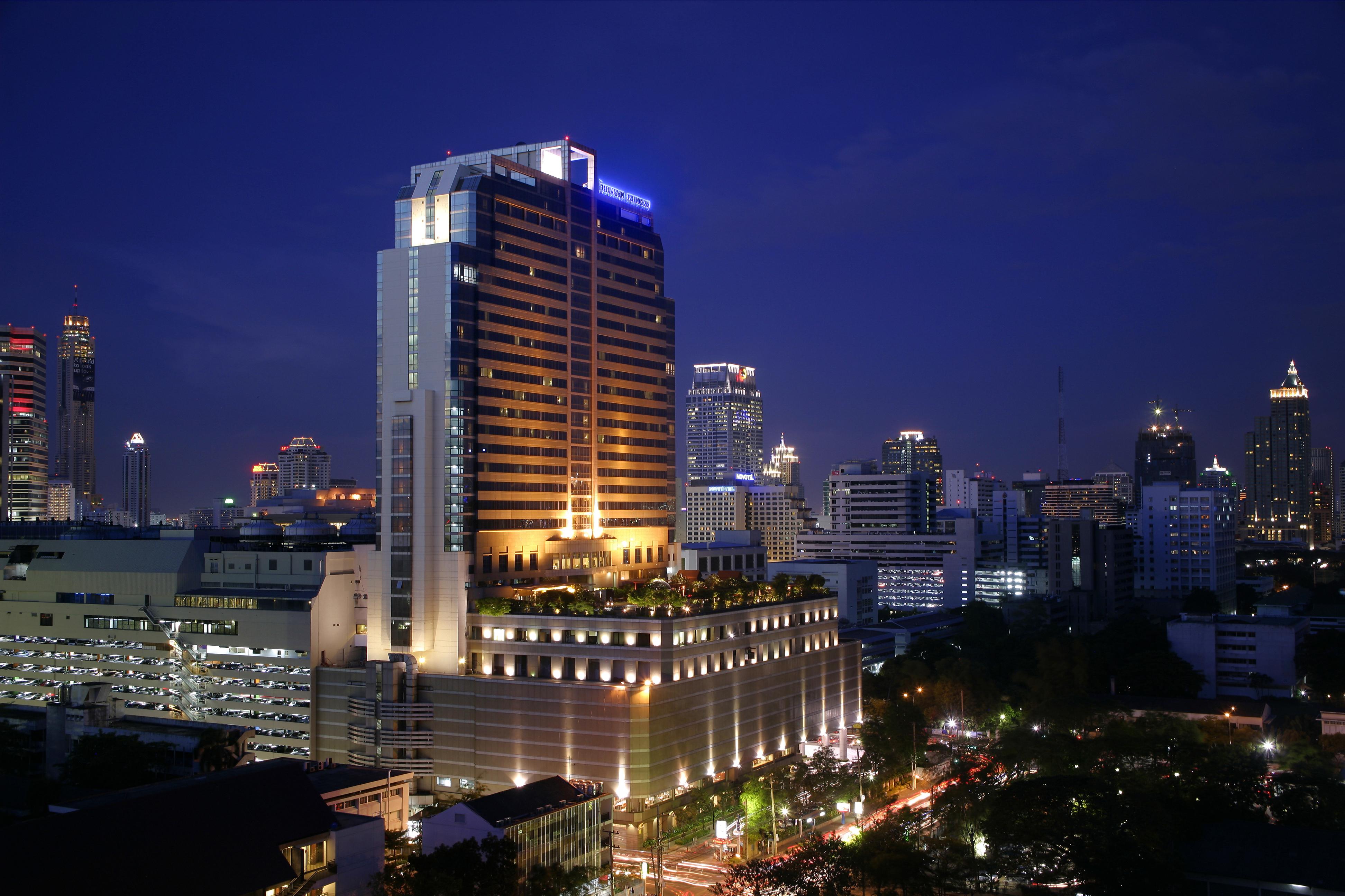 Pathumwan Princess Hotel - Sha Extra Plus Certified Banguecoque Exterior foto The photo shows a cityscape at night, featuring a tall, modern hotel building illuminated with bright lights. The building has a sleek design and is surrounded by a variety of other skyscrapers and urban structures. The street below is bustling with 