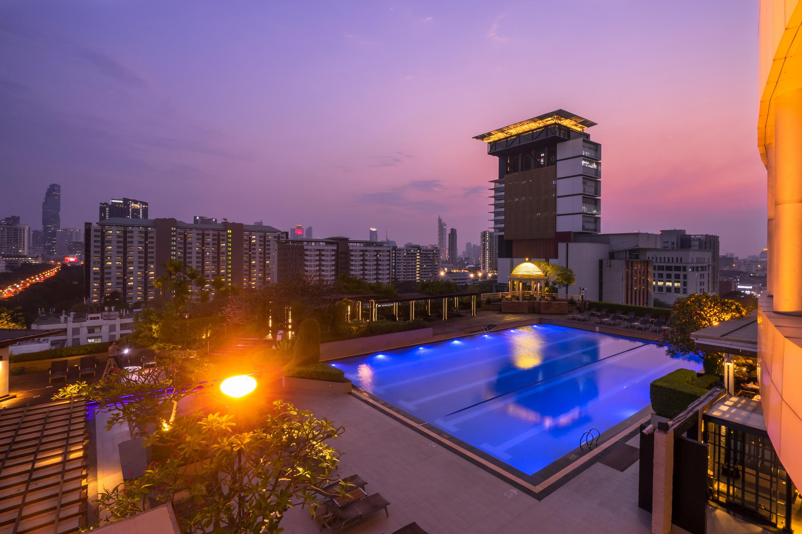 Pathumwan Princess Hotel - Sha Extra Plus Certified Banguecoque Exterior foto The photo depicts a serene rooftop scene featuring a large swimming pool illuminated with blue lighting. Surrounding the pool are lush green trees and landscaping, creating a relaxing atmosphere. In the background, there are modern buildings silhouet
