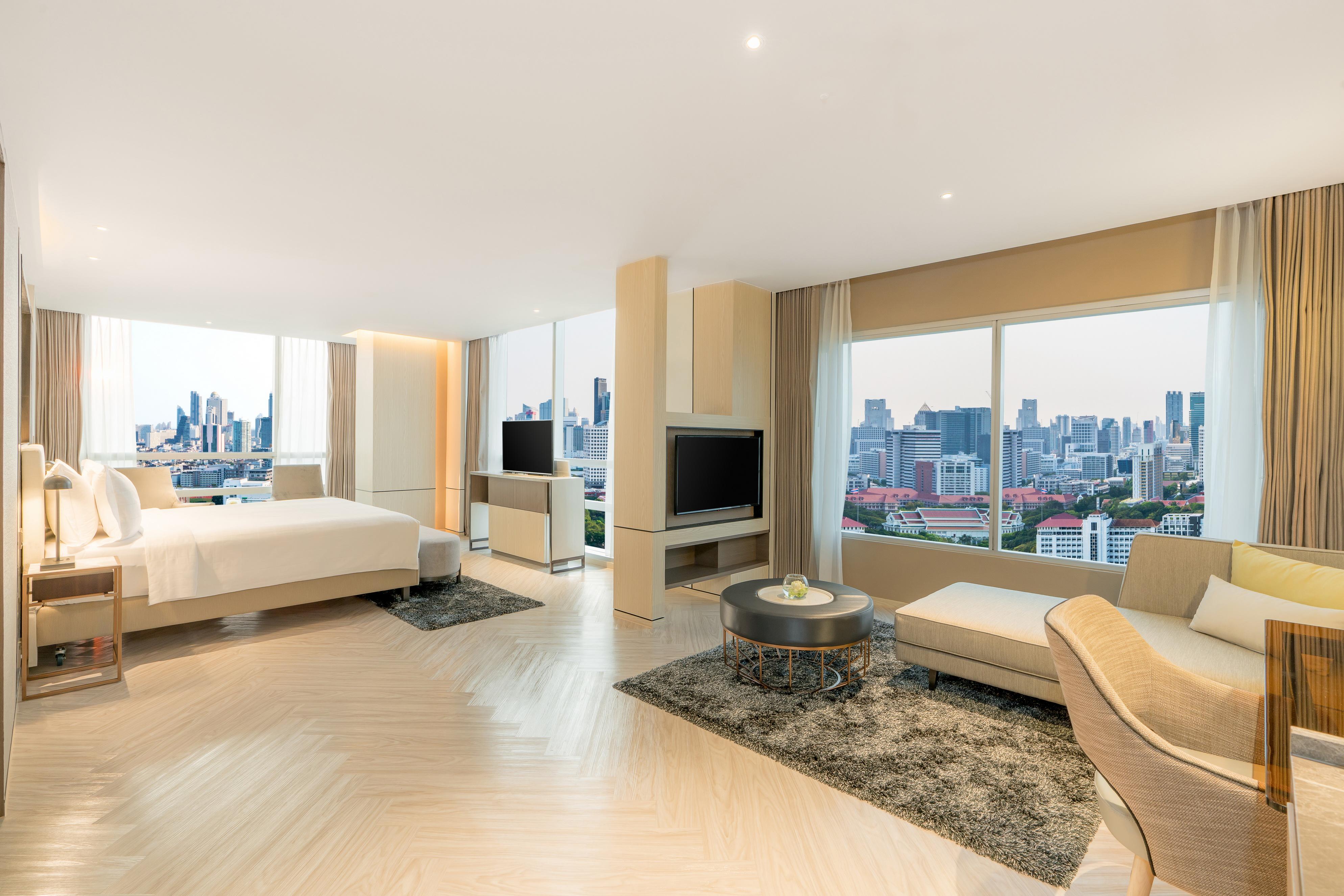 Pathumwan Princess Hotel - Sha Extra Plus Certified Banguecoque Exterior foto The photo shows a modern hotel room featuring a spacious layout. It includes a large bed, a comfortable seating area with a light-colored armchair, and a coffee table on a plush rug. There are large windows that offer a panoramic view of a city skyli