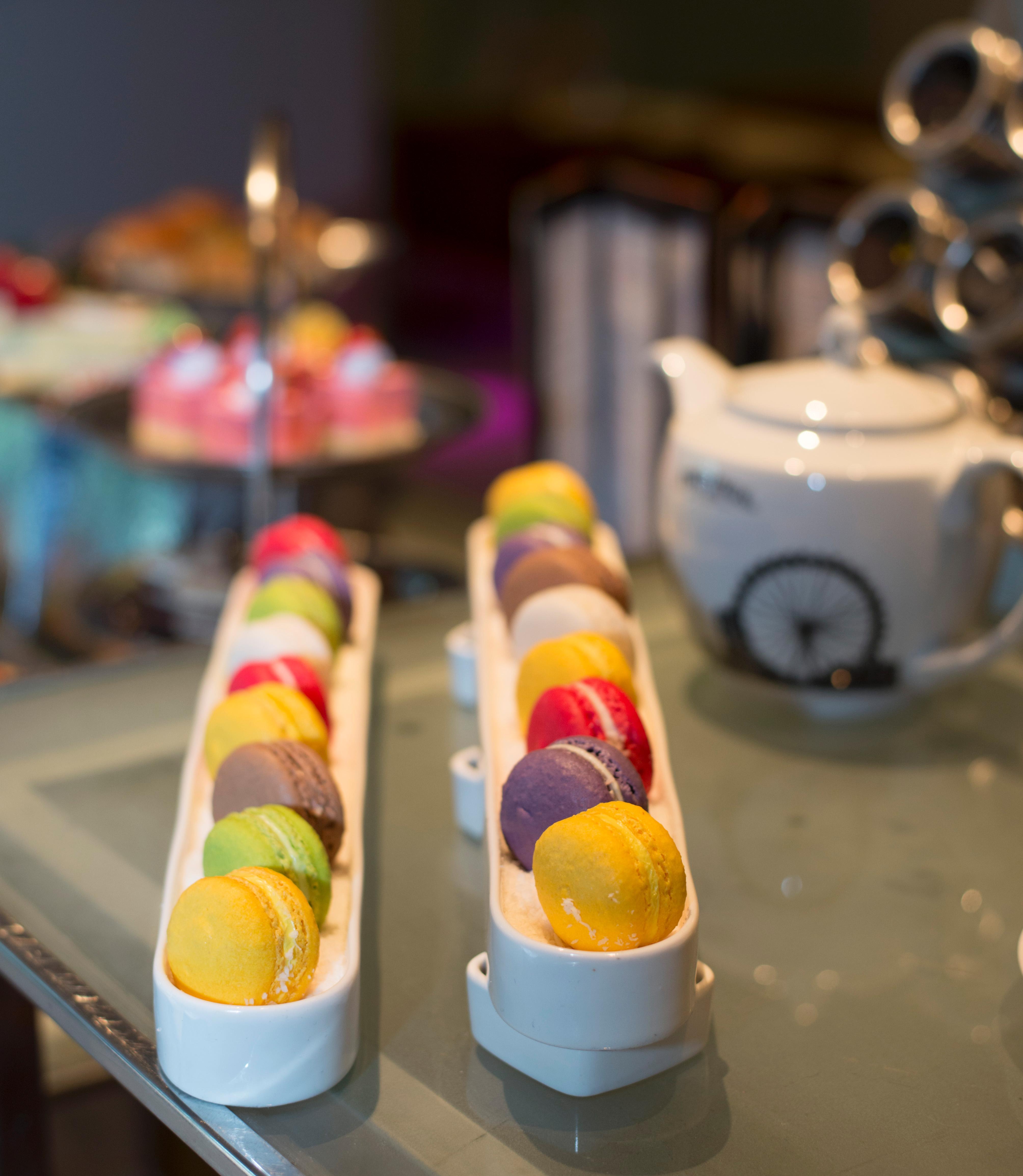 Pathumwan Princess Hotel - Sha Extra Plus Certified Banguecoque Exterior foto The photo showcases an elegant display of macarons arranged in two long, narrow trays. The macarons come in a variety of colors, including yellow, green, pink, purple, and orange, suggesting a range of flavors. In the background, there is a white tea