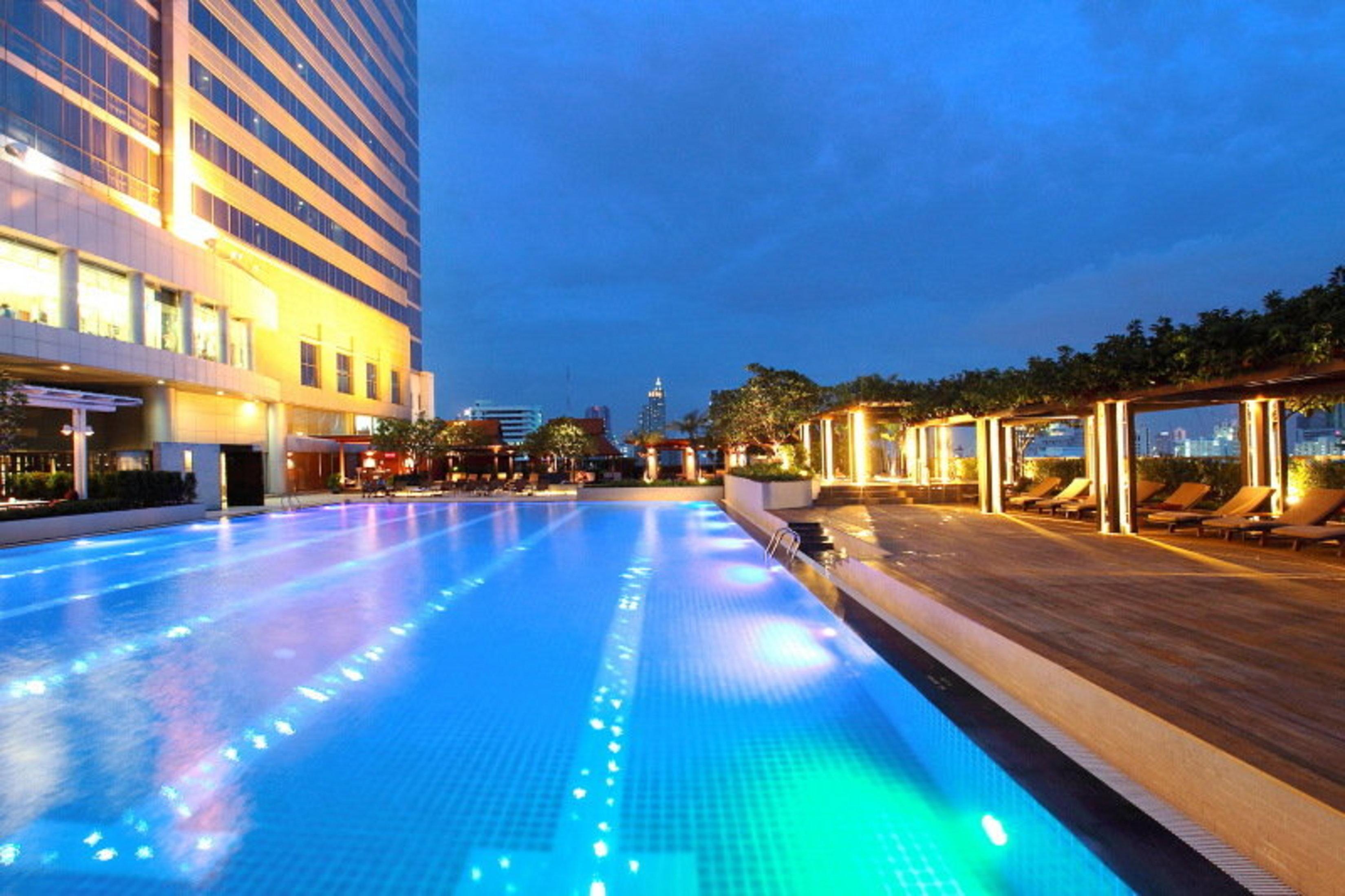 Pathumwan Princess Hotel - Sha Extra Plus Certified Banguecoque Exterior foto The photo showcases a luxurious swimming pool at dusk. The pool is illuminated with colorful lights that create a vibrant atmosphere. Surrounding the pool are lounge areas with wooden decking and lounge chairs, providing a relaxing space for guests. 