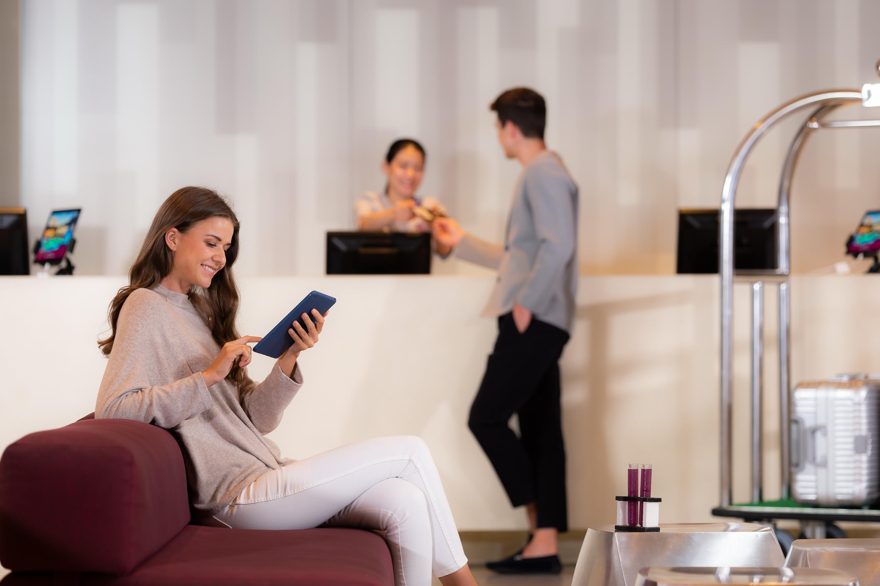 Pathumwan Princess Hotel - Sha Extra Plus Certified Banguecoque Exterior foto The photo shows a modern reception area. In the foreground, a woman is sitting on a maroon sofa, engaged with a tablet. She is wearing a light sweater and white pants. In the background, two people are at the reception desk. One person is a staff mem