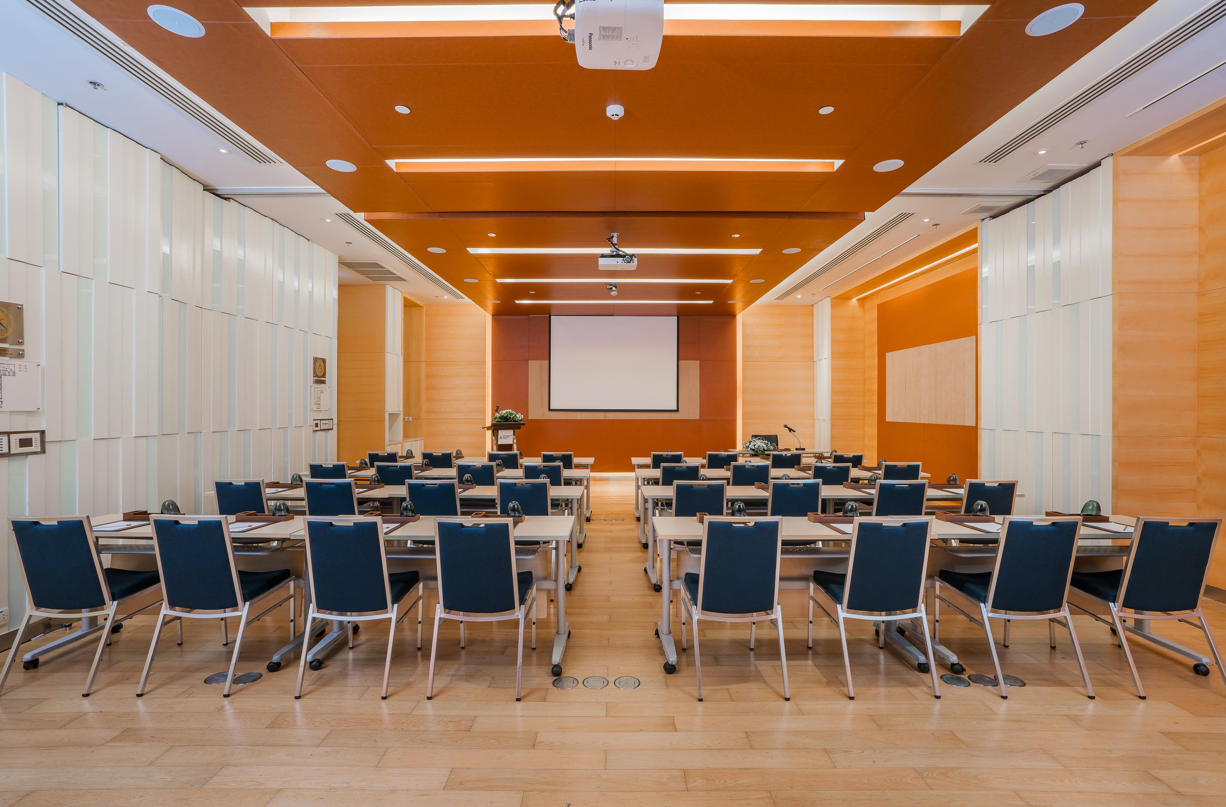 Pathumwan Princess Hotel - Sha Extra Plus Certified Banguecoque Exterior foto The photo shows a modern conference room or seminar space. It features a clean, organized layout with rows of tables and blue chairs arranged facing a large blank screen at the front of the room. The walls have a warm orange hue, and there are bright