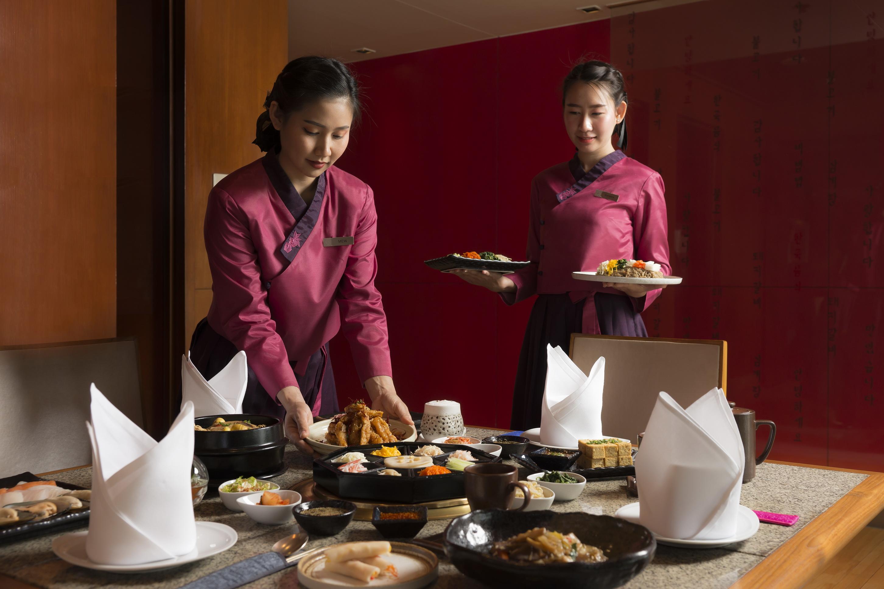Pathumwan Princess Hotel - Sha Extra Plus Certified Banguecoque Exterior foto The photo depicts two women in traditional attire, likely serving food in a restaurant setting. One woman is placing dishes on a table, which is elegantly set with various plates of food. The other woman stands nearby holding a plate of food. The tab
