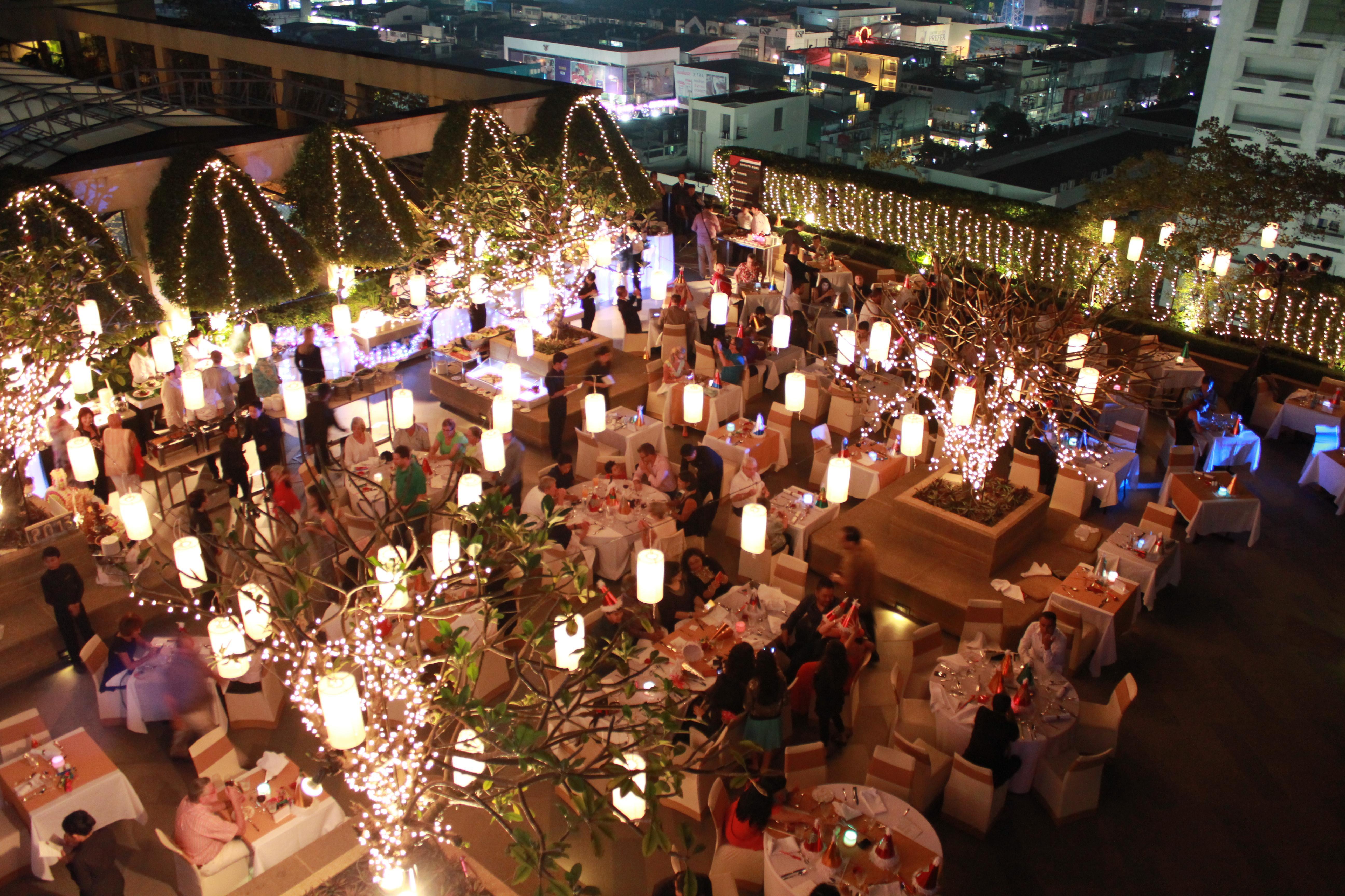 Pathumwan Princess Hotel - Sha Extra Plus Certified Banguecoque Exterior foto The image depicts a spacious outdoor dining area at night, filled with tables arranged for guests. The setting is illuminated by warm, inviting lights, including hanging lanterns and fairy lights draped across trees. Many diners are visible, enjoying
