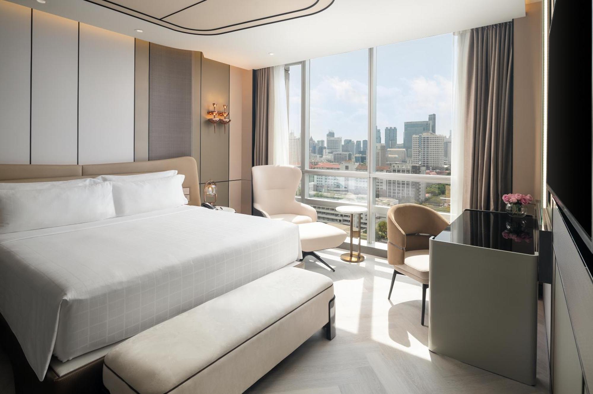 Pathumwan Princess Hotel - Sha Extra Plus Certified Banguecoque Exterior foto The photo shows a modern hotel room featuring a large bed with white linens. There are two stylish chairs and a small table positioned near a large window that offers a view of the city skyline. A sleek desk is also present, with flowers placed on it