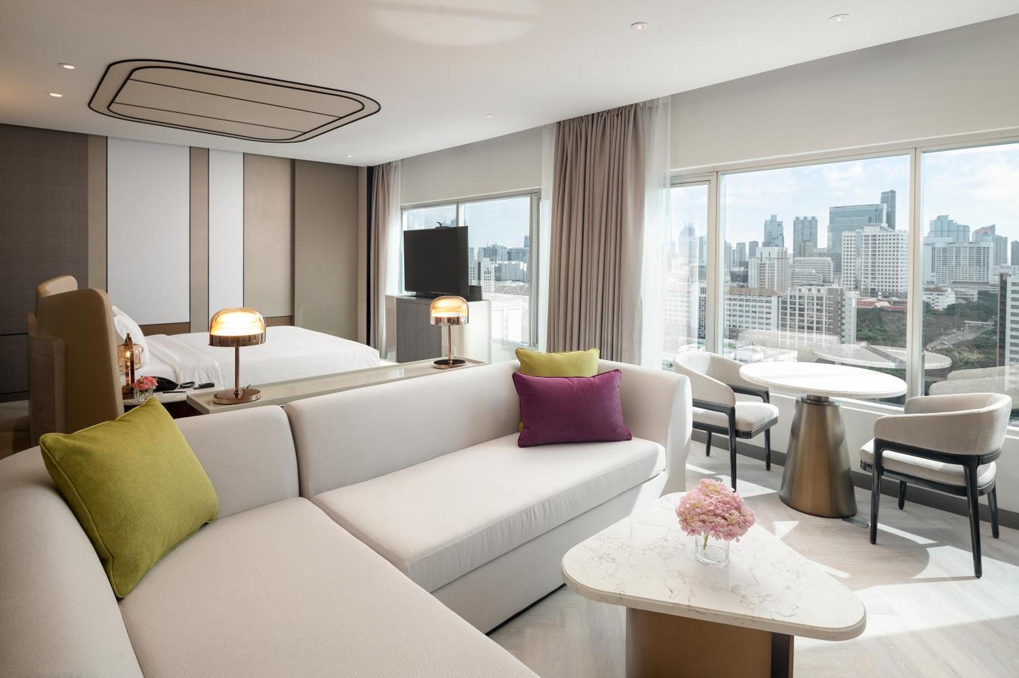 Pathumwan Princess Hotel - Sha Extra Plus Certified Banguecoque Exterior foto The photo showcases a modern hotel room featuring a stylish interior design. There is a large, comfortable seating area with a white sofa adorned with colorful cushions in green and purple. Nearby, a small round table with a floral centerpiece stands