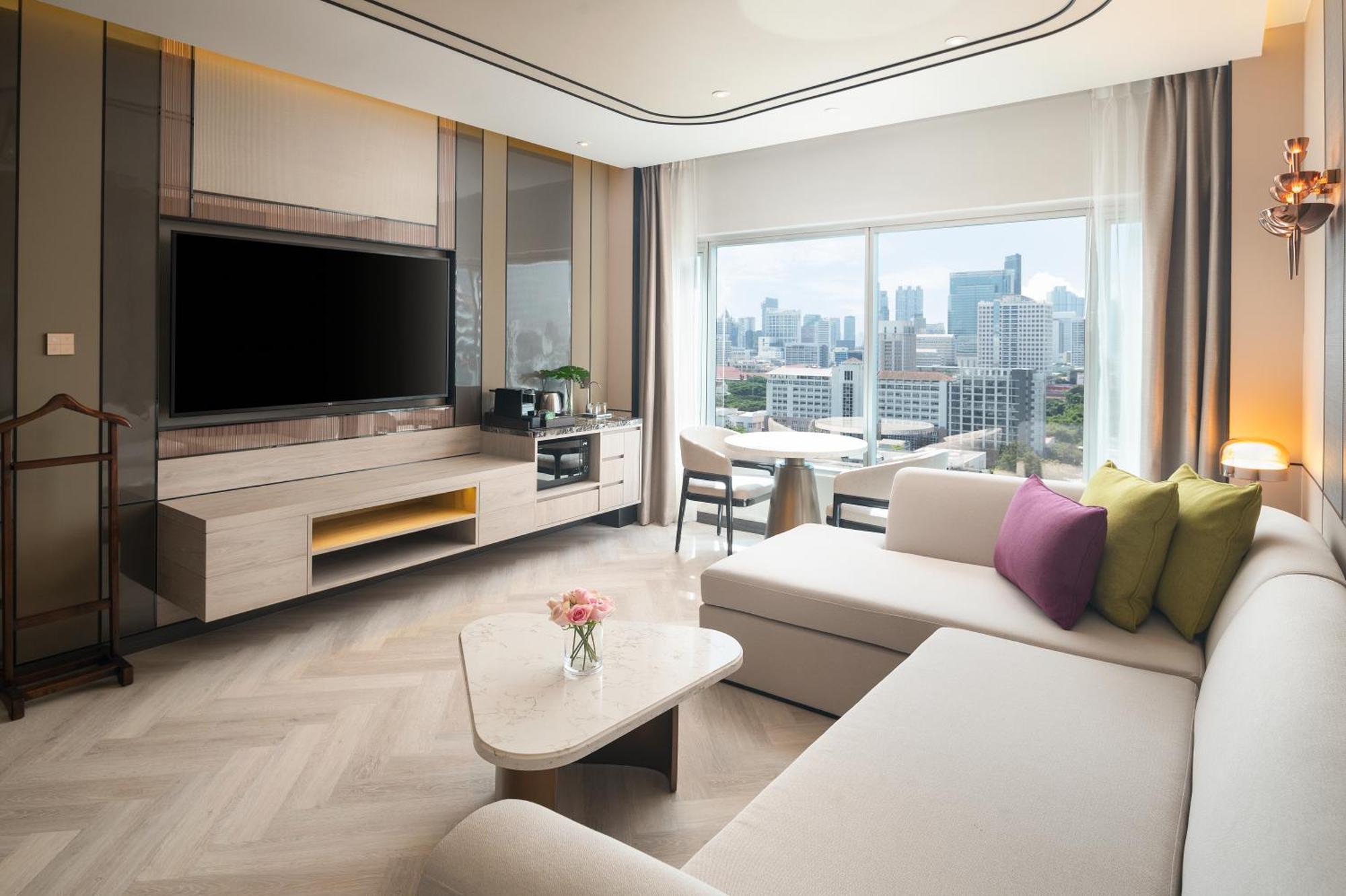 Pathumwan Princess Hotel - Sha Extra Plus Certified Banguecoque Exterior foto The photo shows a modern living room with a large window offering a view of a city skyline. The room features a light-colored sofa in an L-shape, complemented by colorful cushions in shades of green and purple. A sleek coffee table sits in front of t