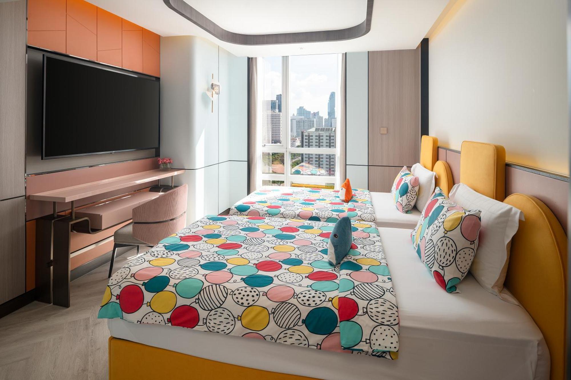 Pathumwan Princess Hotel - Sha Extra Plus Certified Banguecoque Exterior foto The photo shows a brightly decorated hotel room featuring two beds with colorful, polka-dot patterned bedspreads. The beds have contrasting headboards in orange and yellow. There is a modern desk and a television mounted on the wall opposite the beds