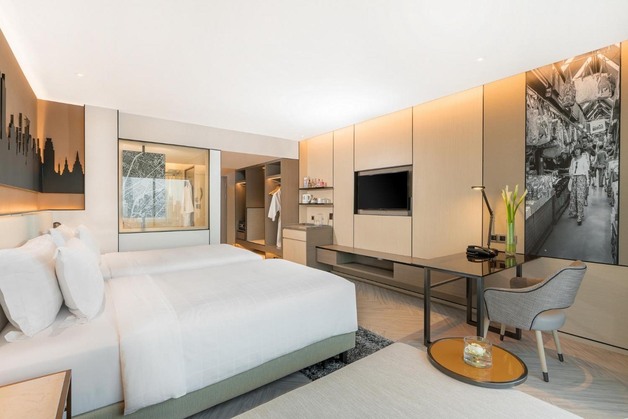 Pathumwan Princess Hotel - Sha Extra Plus Certified Banguecoque Quarto foto The photo shows a modern hotel room featuring a minimalist design. There are two beds with white bedding, positioned against one wall. A stylish desk and chair are situated near a large window, which allows natural light to fill the space. A flat-scr