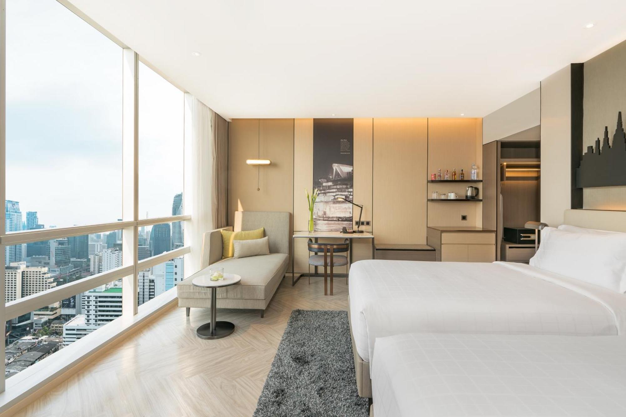 Pathumwan Princess Hotel - Sha Extra Plus Certified Banguecoque Quarto foto The photo shows a modern and stylish hotel room. It features two beds, a small lounge area with a sofa, and a table. Large windows allow for plenty of natural light and offer a view of the cityscape outside. The interior has a contemporary design wit