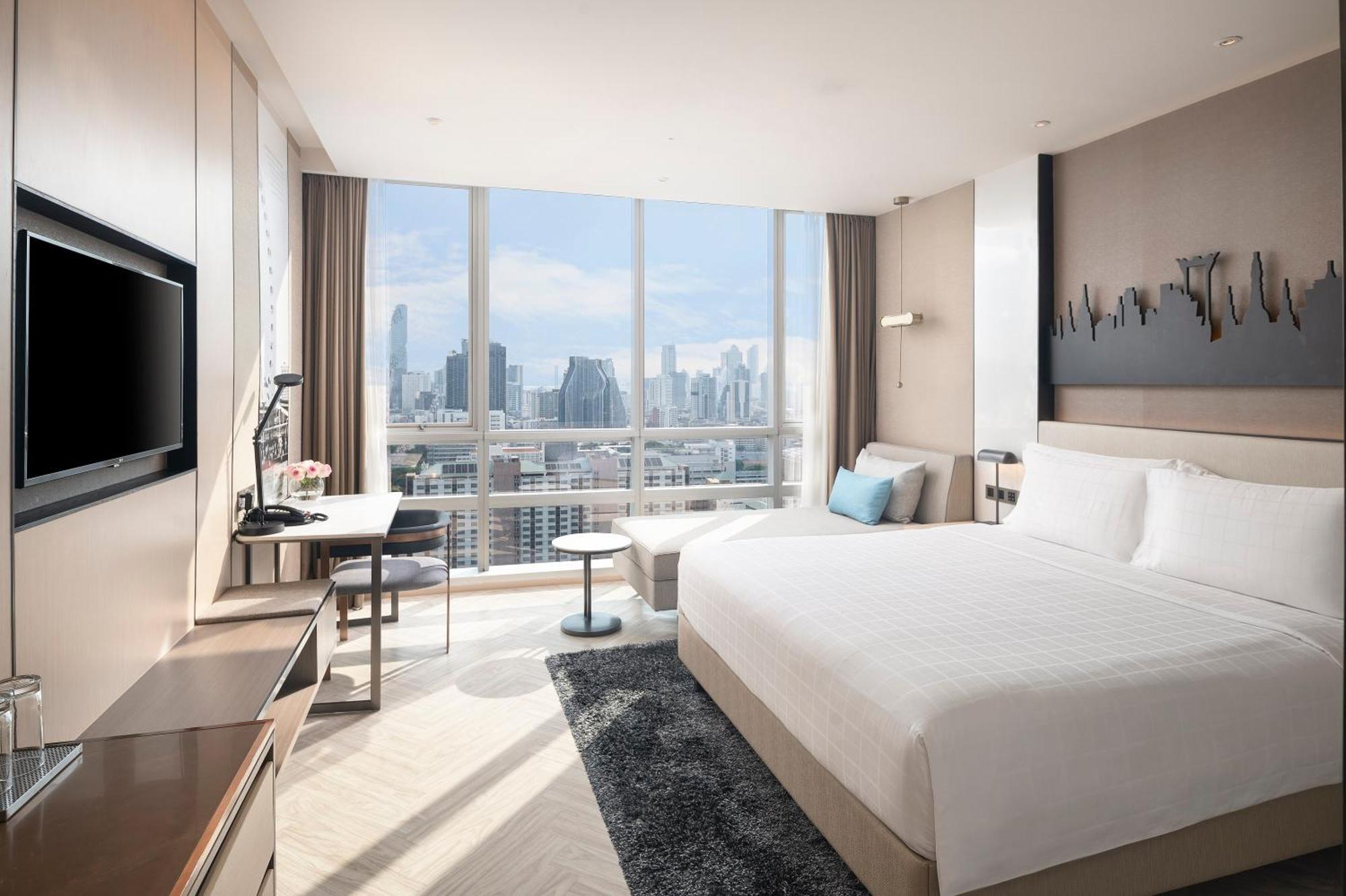 Pathumwan Princess Hotel - Sha Extra Plus Certified Banguecoque Exterior foto The photo shows a modern hotel room with large windows that provide a view of the city skyline. The room features a neatly made king-sized bed, a bedside table, and a chair in the corner. There's a desk with a lamp and a flat-screen TV mounted on the