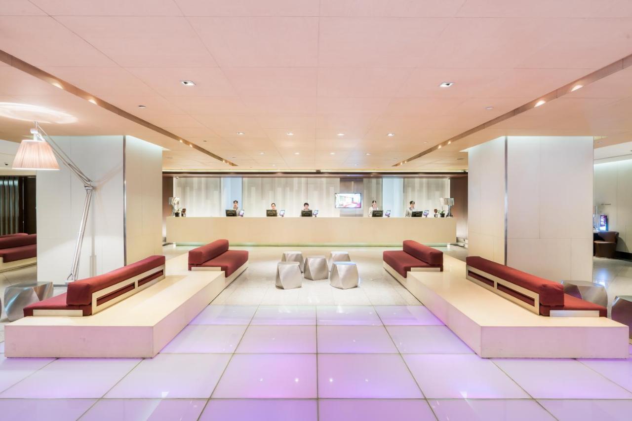 Pathumwan Princess Hotel - Sha Extra Plus Certified Banguecoque Exterior foto The photo shows a modern reception area, likely in a hotel or upscale building. The space features a light-colored floor with a subtle purple glow, which adds a contemporary touch. There are several sleek, minimalist benches arranged around the area,