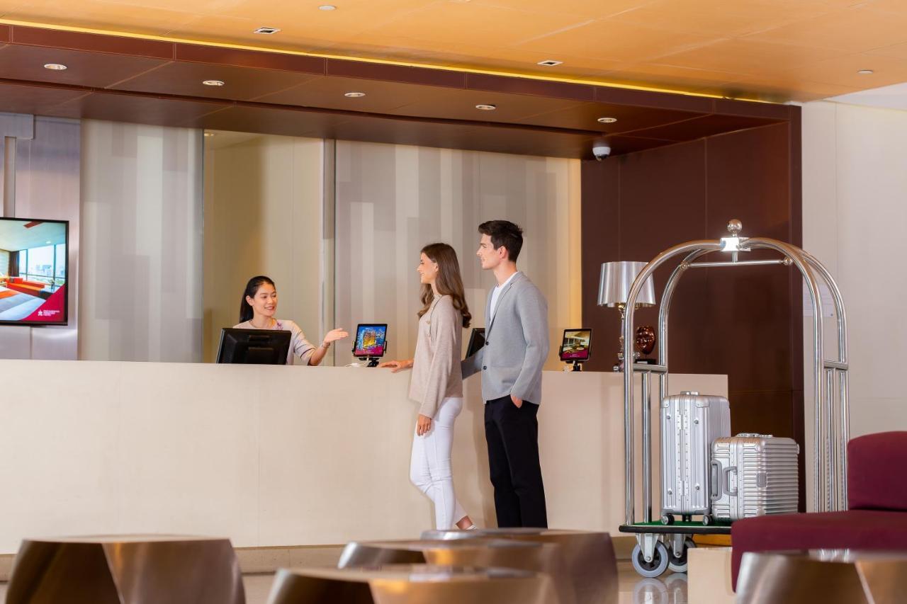 Pathumwan Princess Hotel - Sha Extra Plus Certified Banguecoque Exterior foto The photo shows a hotel reception area. In the foreground, a receptionist is interacting with a couple. The receptionist, dressed in professional attire, is standing behind a sleek front desk, which has a digital screen displaying information. The co