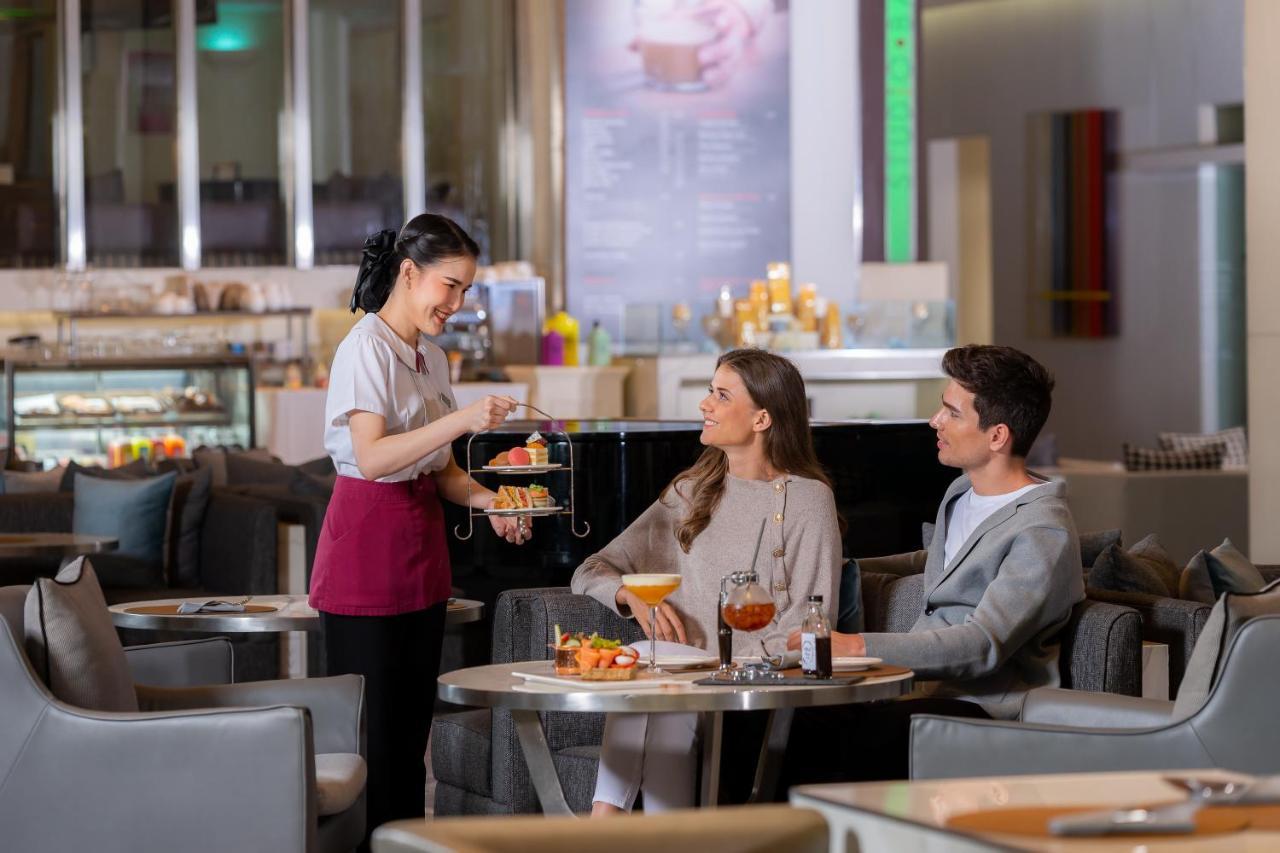 Pathumwan Princess Hotel - Sha Extra Plus Certified Banguecoque Exterior foto The photo shows a scene in a stylish cafe or restaurant. A waitress is serving a dessert stand to two guests who are seated at a table. The waitress is dressed in a uniform and appears to be presenting a selection of desserts. The two guests, a man a