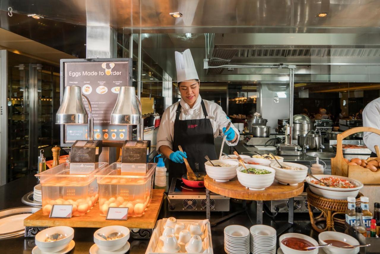 Pathumwan Princess Hotel - Sha Extra Plus Certified Banguecoque Exterior foto The photo shows a chef working in a bustling kitchen environment. He is wearing a white chef's hat and an apron, along with blue gloves for hygiene. In front of him, there are several bowls and ingredients for preparing dishes. A display sign indicat