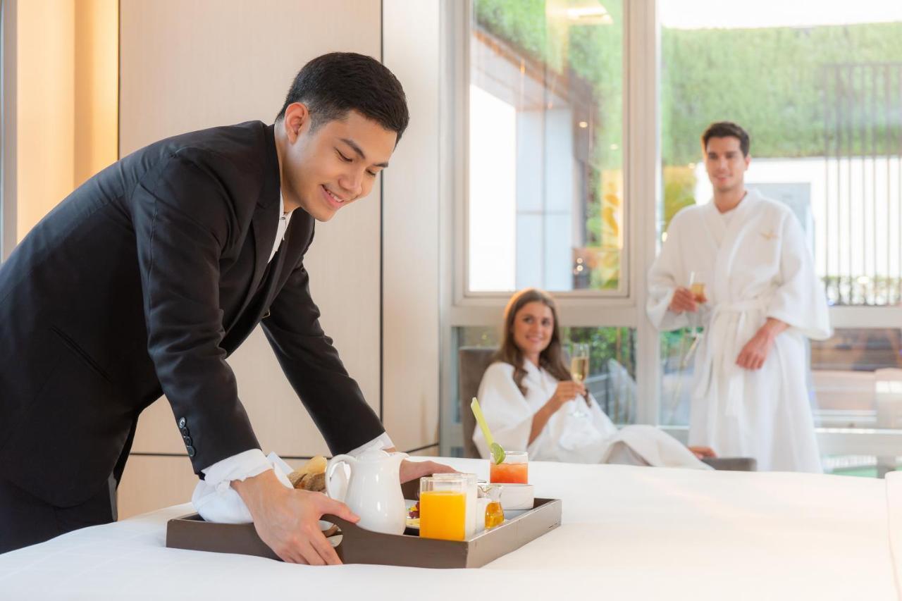 Pathumwan Princess Hotel - Sha Extra Plus Certified Banguecoque Exterior foto The photo depicts a luxurious hotel setting. A server, dressed in formal attire, is placing a tray with beverages, including orange juice and a teapot, on a bed. In the background, a woman is sitting on the bed in a robe, looking pleased, while a man
